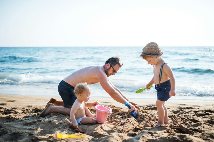 Vater spielt mit zwei kleinen Kindern im Sand am Strand, während das Wasser plätschert.