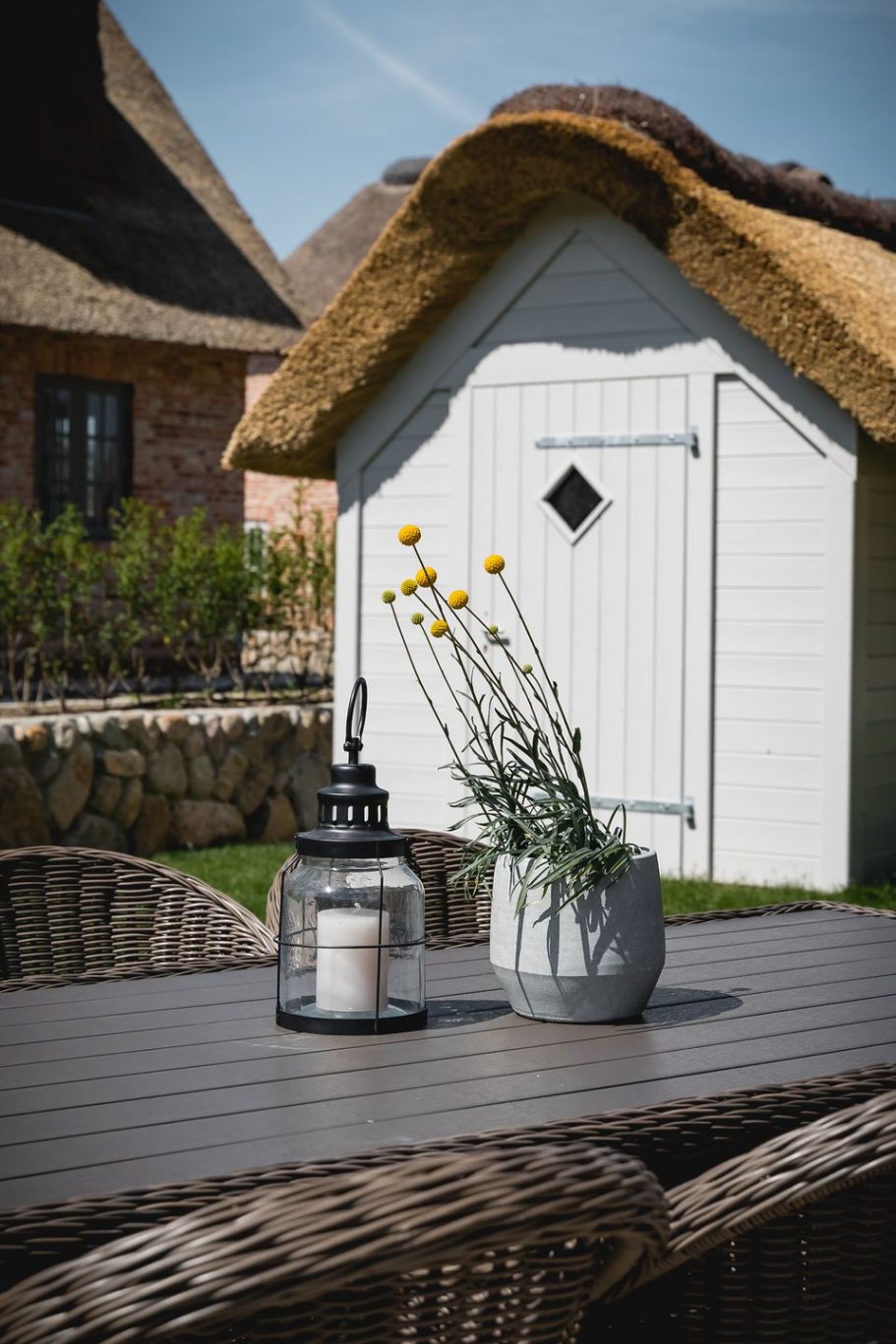 Ein Tisch mit Pflanzen und Laterne vor einem weißen Gartenhaus mit Reetdach.