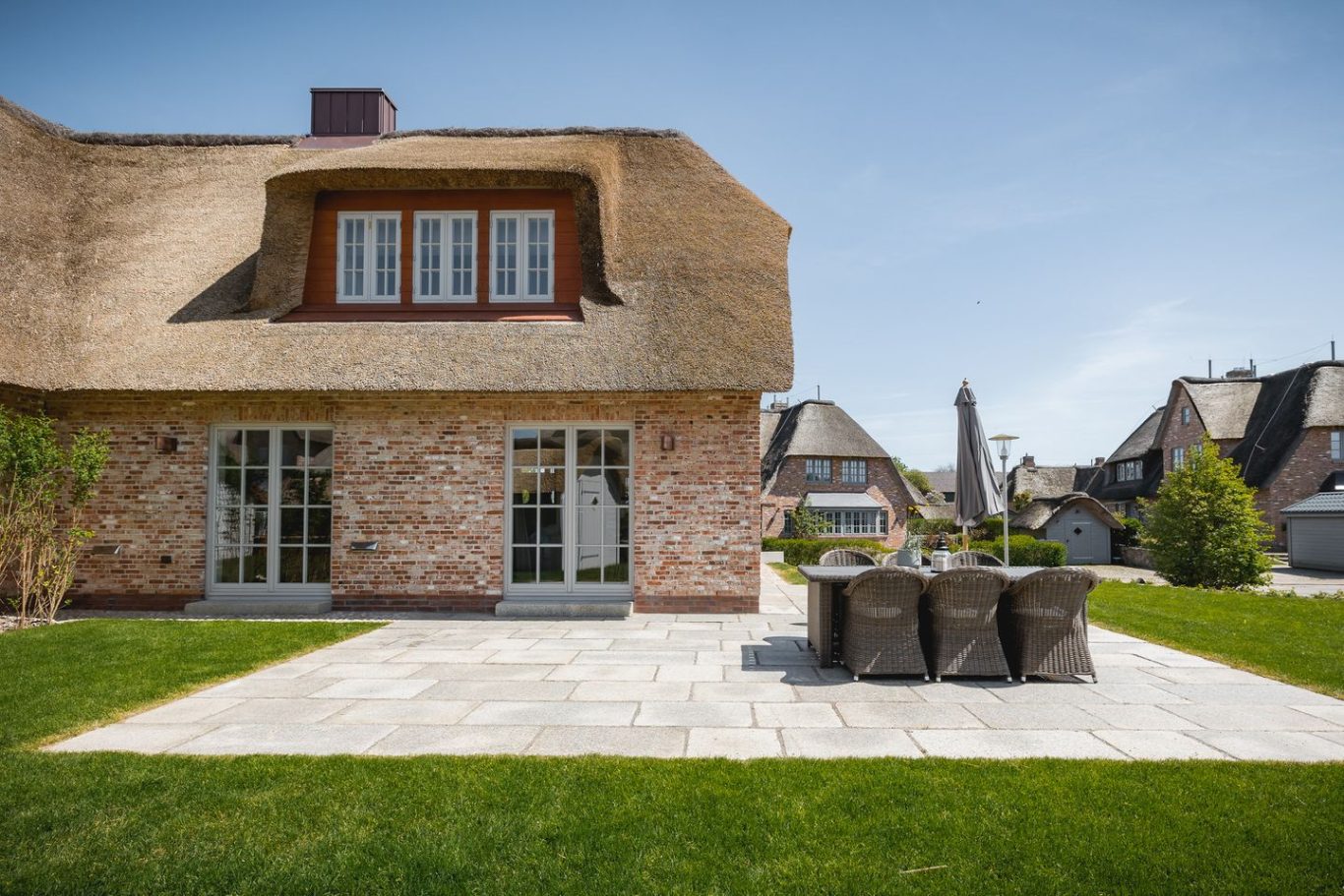 Haus mit Reetdach, Terrasse und Gartenmöbeln in einer sonnigen Umgebung.