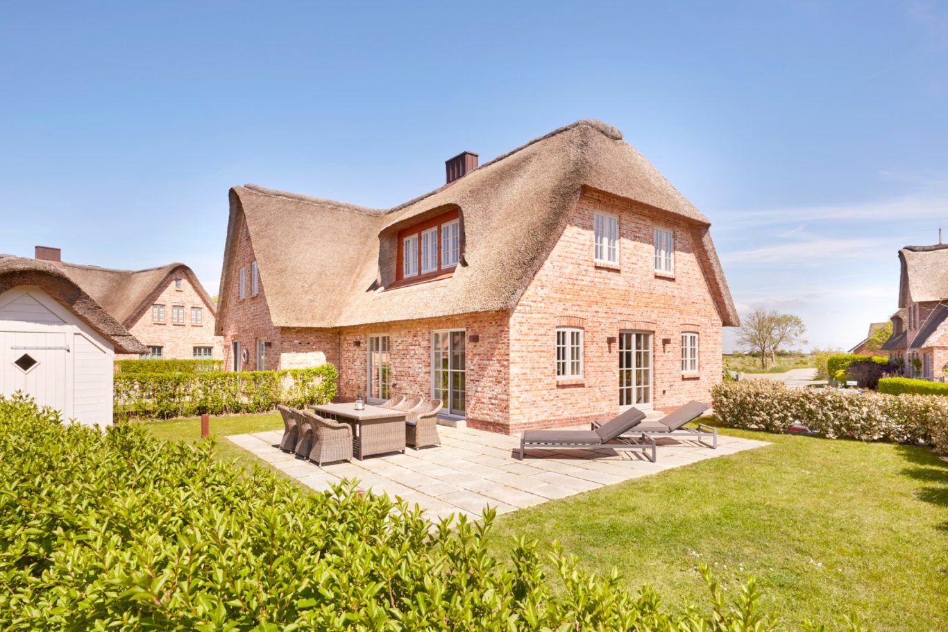 Süd-West-Terrasse Haus mit Reetdach und Garten, umgeben von grünen Hecken und klarem blauen Himmel.