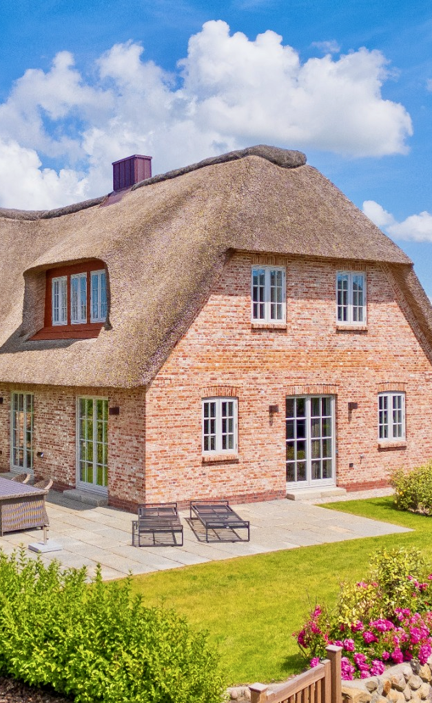 Ferienhaus mit Reetdach, Garten und Terrasse unter blauem Himmel.